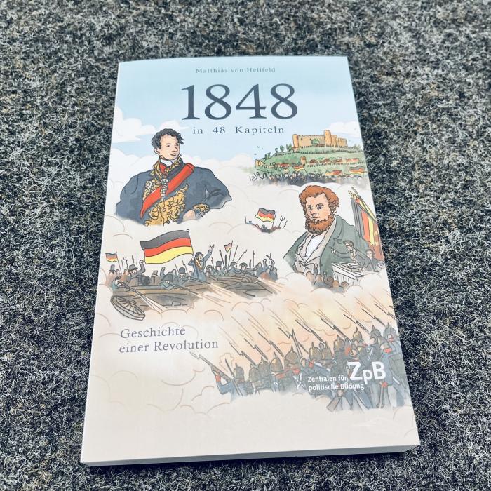 Zu sehen ist der Umschlag des Buches „1848 in 48 Kapiteln - Geschichte einer Revolution“ mit illustrierten Bildern der historischen Persönlichkeiten sowie der Revolutionärs Massen mit Deutschlandfahnen.
