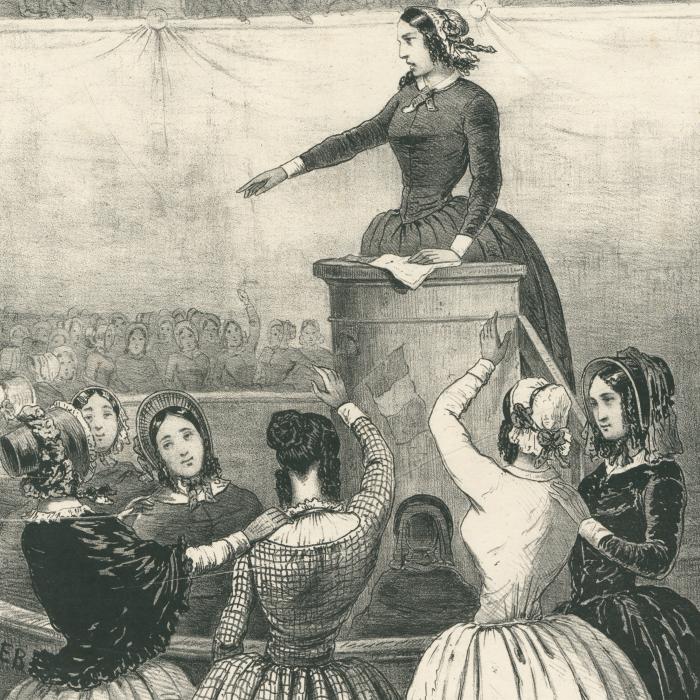  Eine historische Zeichnung. Die Sitzung eines Frauenclubs. Eine Frau steht an einem Rednerpult, im Publikum sind weitere Frauen zu sehen.