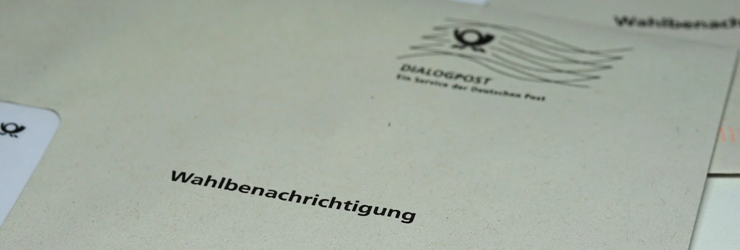 Auf dem Bild ist ein Umschlag mit der Aufschrift "Wahlbenachrichtigung" in klarer, schwarzer Schrift zentral abgebildet. Der Umschlag liegt auf einer Oberfläche neben anderen Papieren.